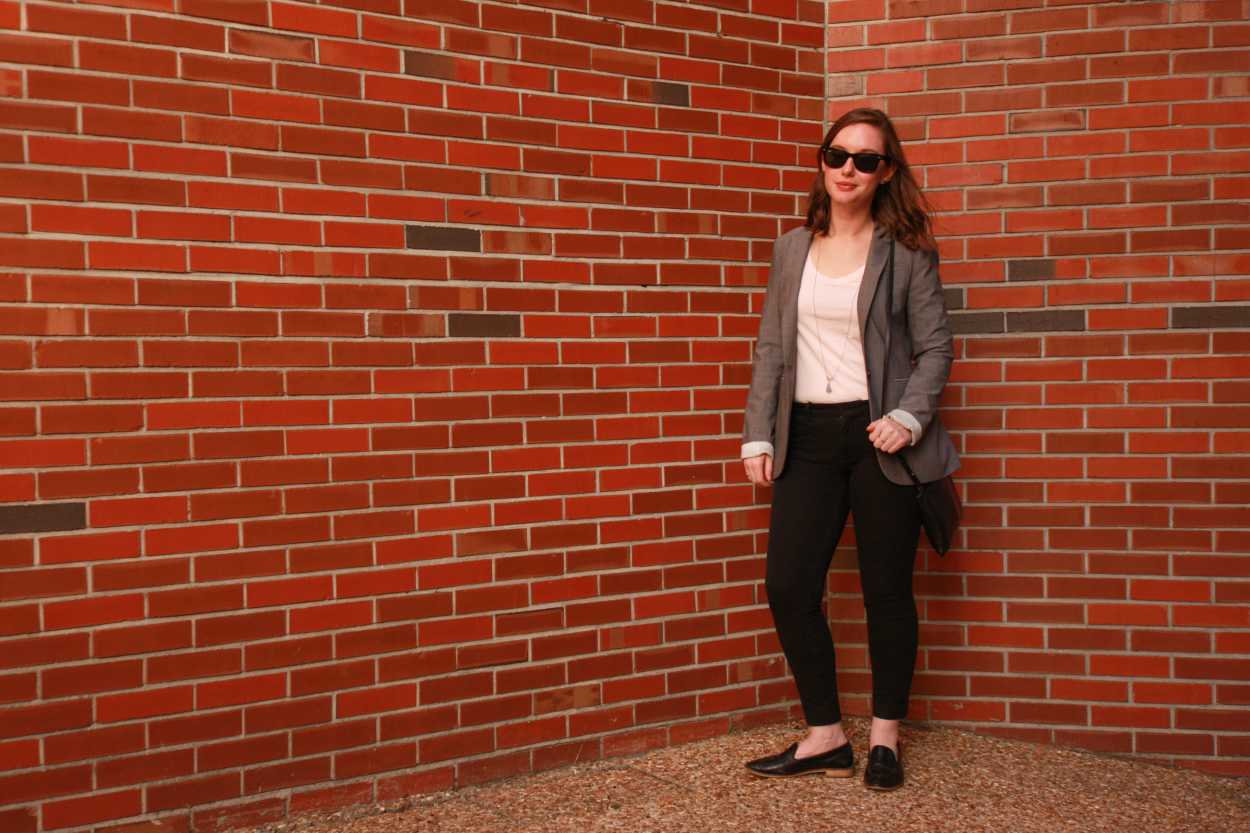 Alyssa wears a blush tee, black pants, black loafers, and a grey blazer. She is by a brick wall, and smiling at the camera.