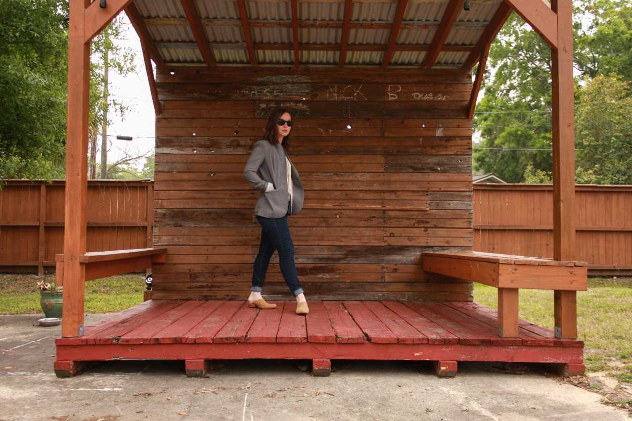 Alyssa wears a white tunic tucked into blue jeans with tan mules and a grey blazer. She is standing on a stage made from wooden pallets and turned halfway toward the camera