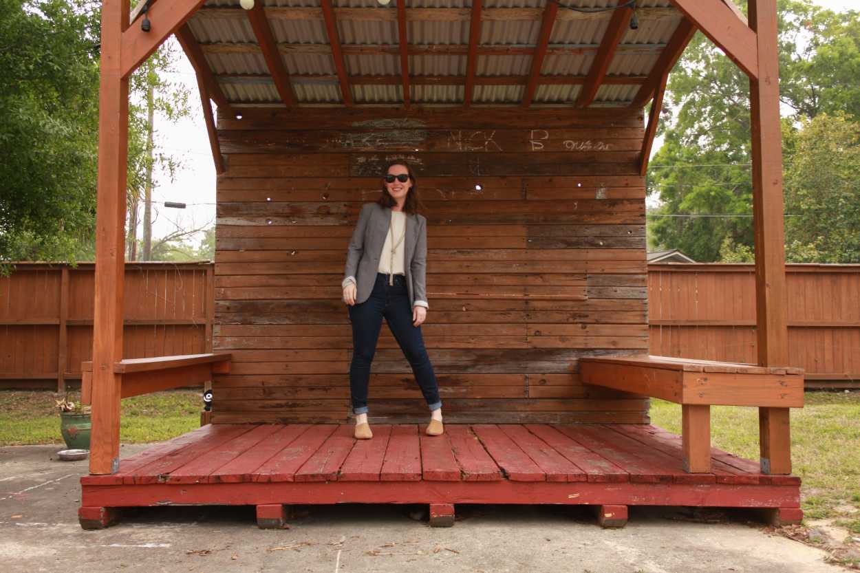 Alyssa wears a white tunic tucked into blue jeans with tan mules and a grey blazer. She is standing on a stage made from wooden pallets and smiling at the camera