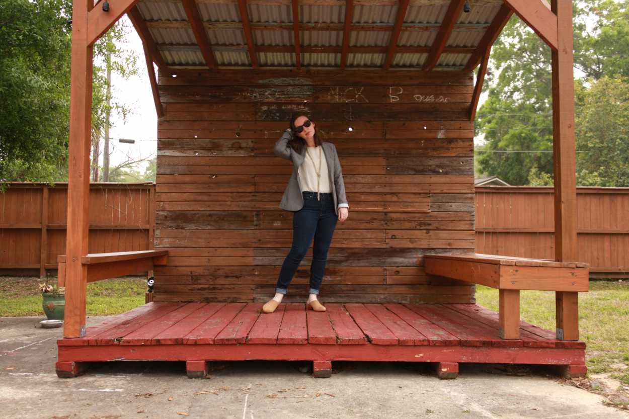 Alyssa wears a white tunic tucked into blue jeans with tan mules and a grey blazer. She is standing on a stage made from wooden pallets and running her hand through her hair