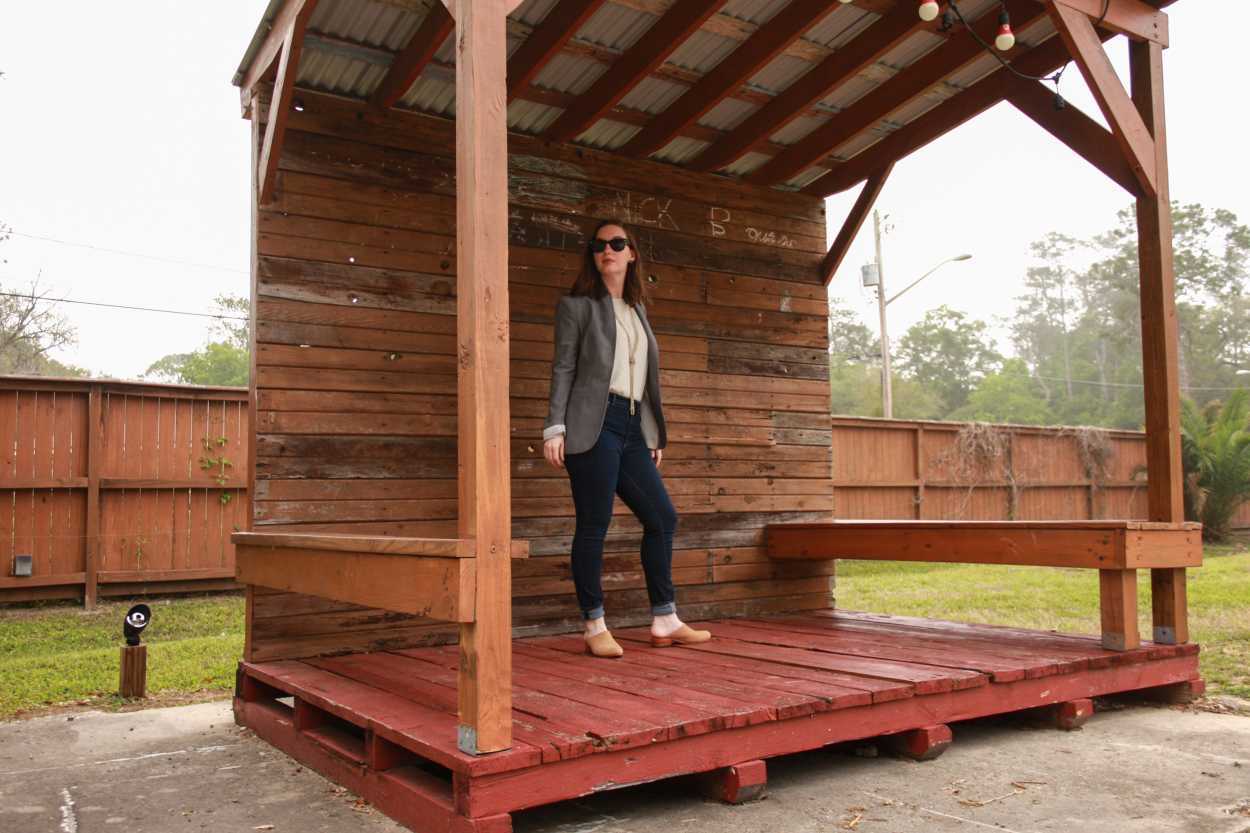Alyssa wears a white tunic tucked into blue jeans with tan mules and a grey blazer. She is standing on a stage made from wooden pallets