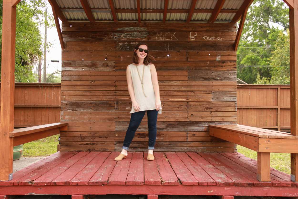 Alyssa wears a white tunic with blue jeans and tan mules. She is standing on a stage made from wooden pallets and smiling