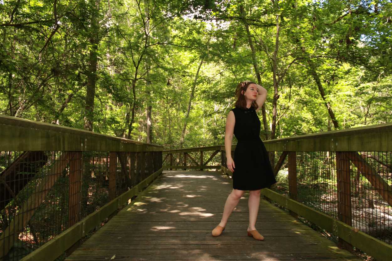 Alyssa wears a black shirtdress with nude mules and stands on a bridge, running her hands through her hair