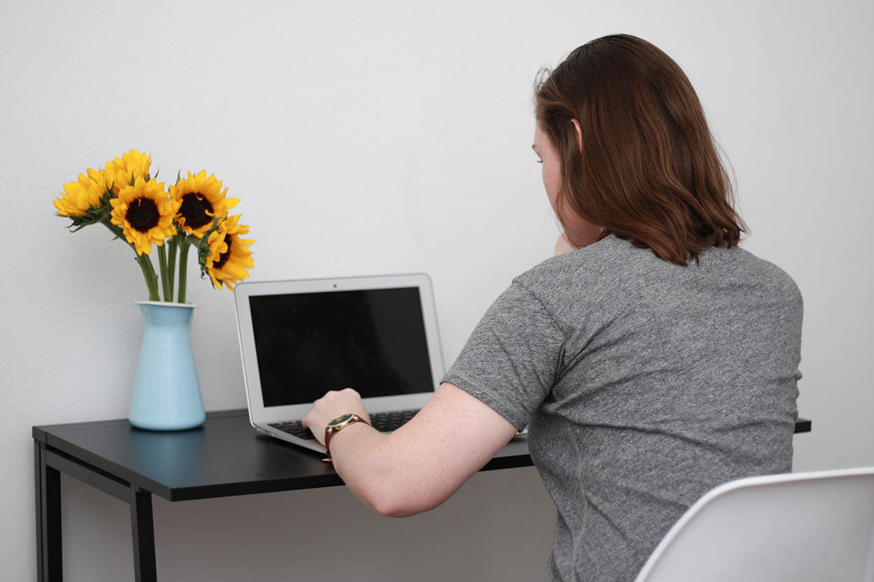 Alyssa sits at a computer set on a desk with sunflowers in a vase