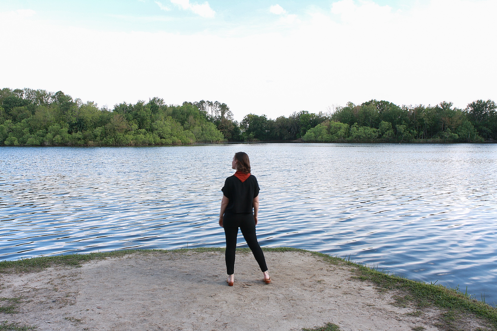 Alyssa is wearing a black silk tee, black pants, tan mules, and a rust bandana. She is standing in front of a lake and facing away from the camera.