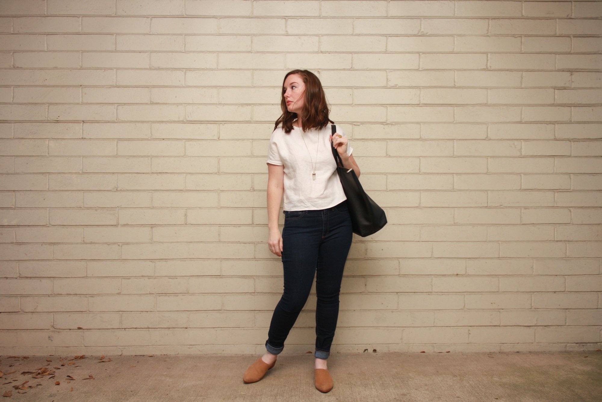 Alyssa wears a homemade white linen tee, skinny blue jeans, and nude mules and carries a tote bag
