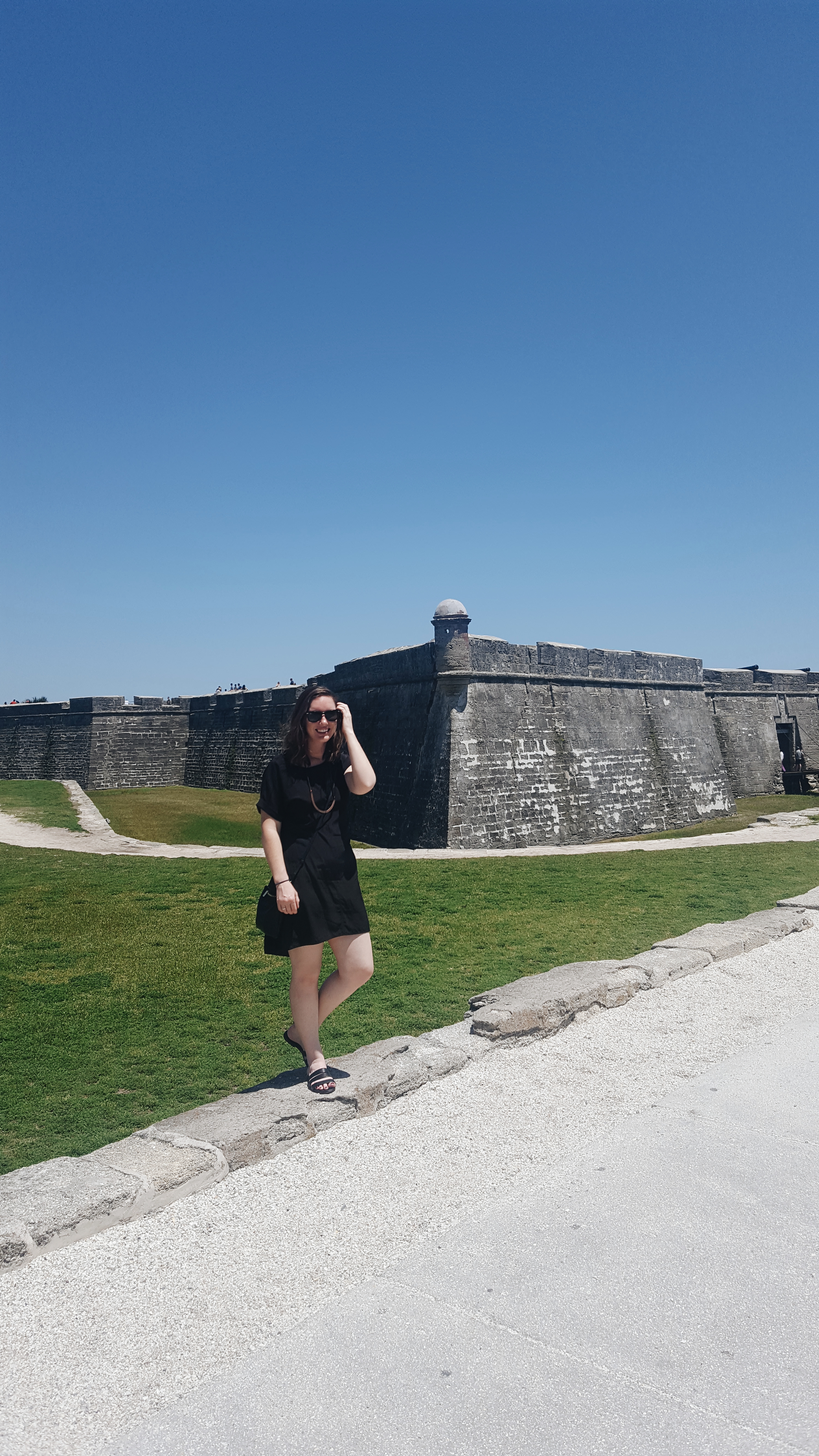 Alyssa stands in front of the St. Augustine fort in a black dress and sandals