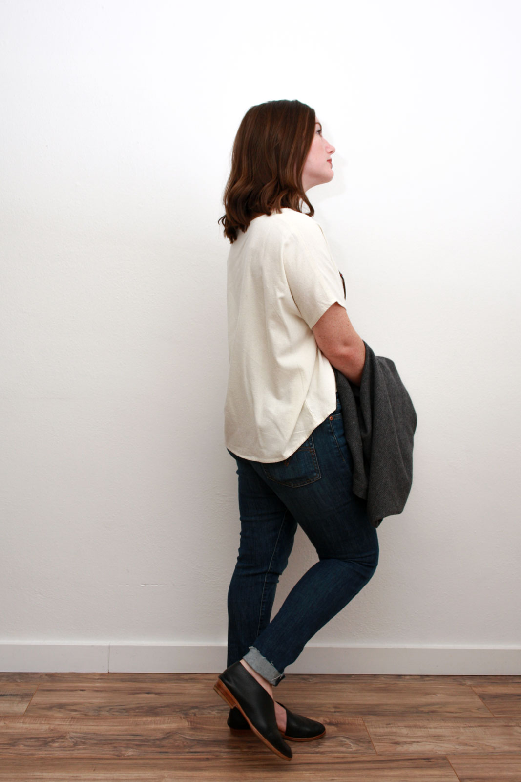 Alyssa wears a cream silk top, blue skinny jeans, and black asymmetrical flats while carrying a grey sweater