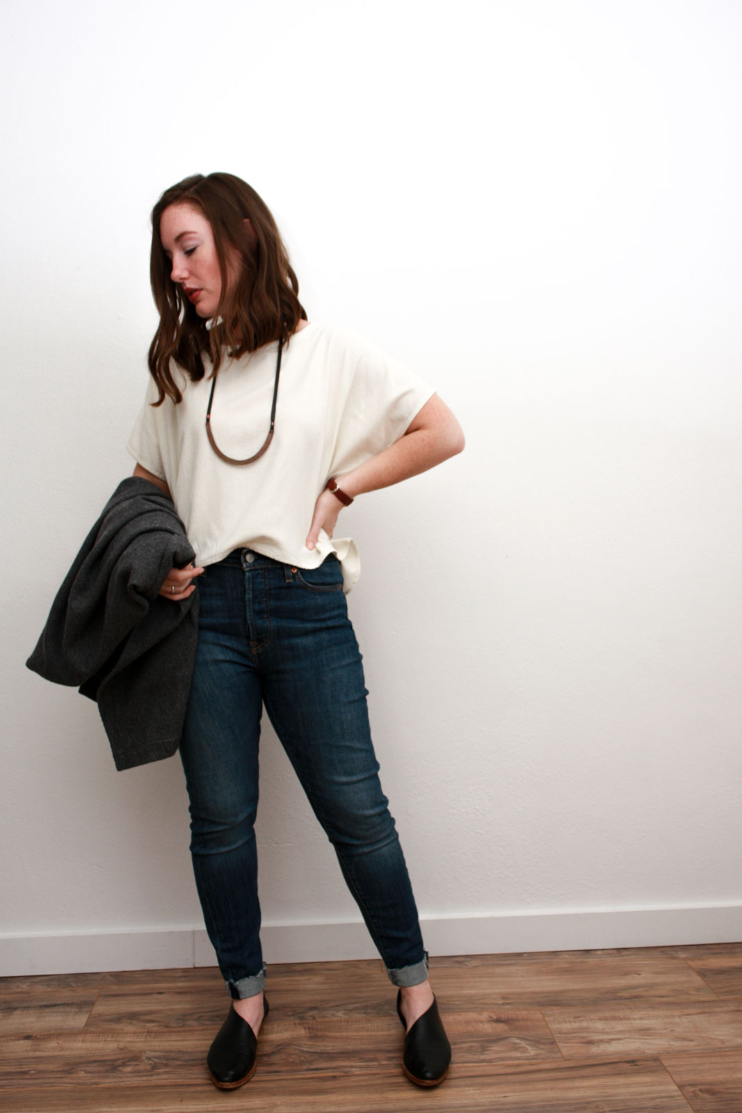 Alyssa wears a cream silk top, blue skinny jeans, and black asymmetrical flats while carrying a grey sweater