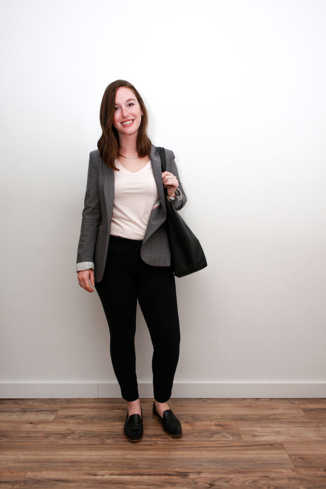 Alyssa wears a pink top, black pants, grey blazer, black loafers and she carries a black tote