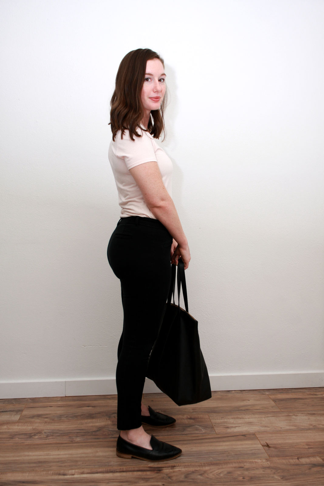 Alyssa wears a pink top, black pants, black loafers, and carries a black tote