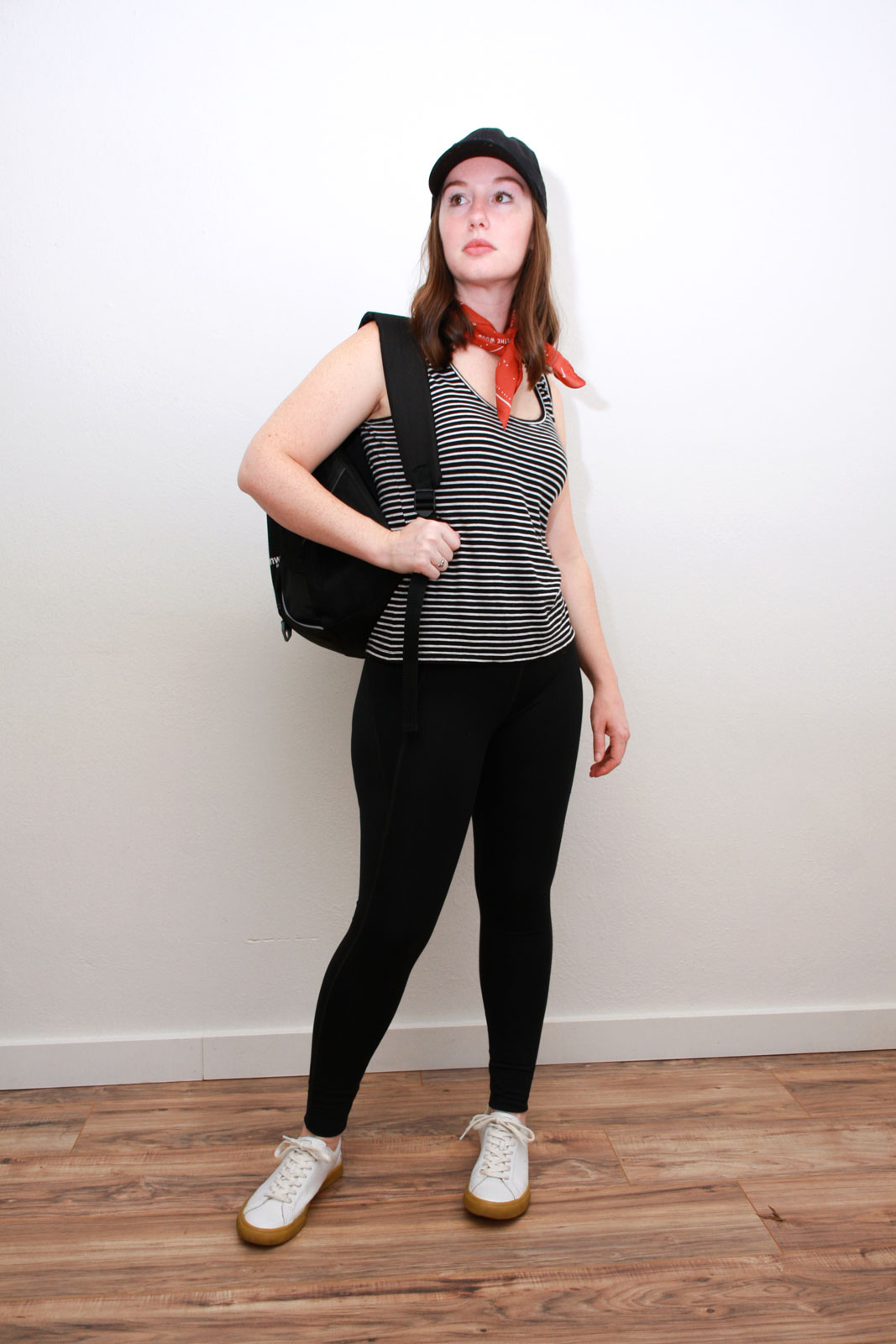Alyssa wears an athleisure outfit of a striped tank, red bandana, black leggings, white sneakers, and black baseball cap. She is carrying a backpack and looking near the camera.