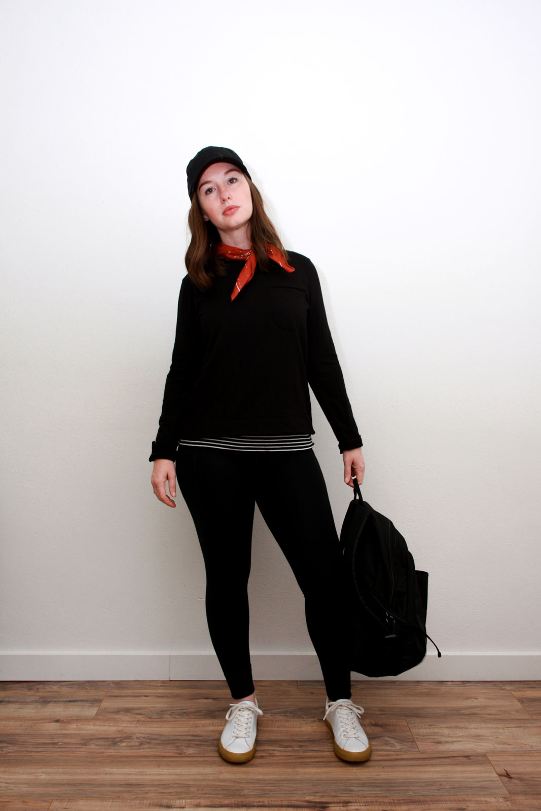 Alyssa wears an athleisure outfit of a striped tank under a black sweatshirt, black leggings, white sneakers, and black baseball cap. She is carrying a backpack and looking at the camera.
