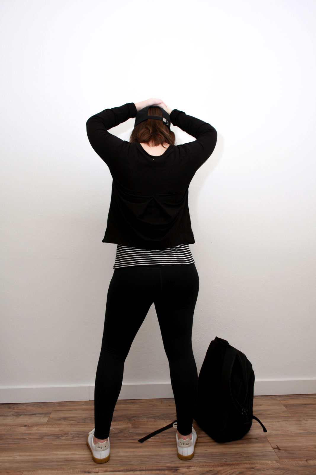 Alyssa wears an athleisure outfit of a striped tank under a black sweatshirt, black leggings, white sneakers, and black baseball cap. She is facing away from the camera