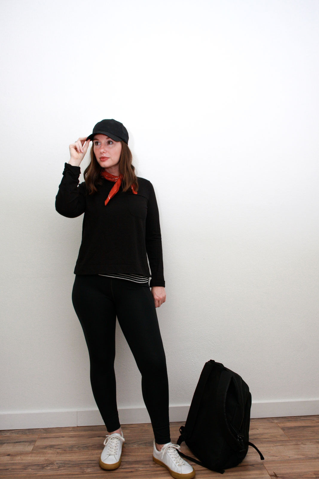 Alyssa wears an athleisure outfit of a striped tank under a black sweatshirt, black leggings, white sneakers, and black baseball cap. She is touching her cap and looking at from the camera
