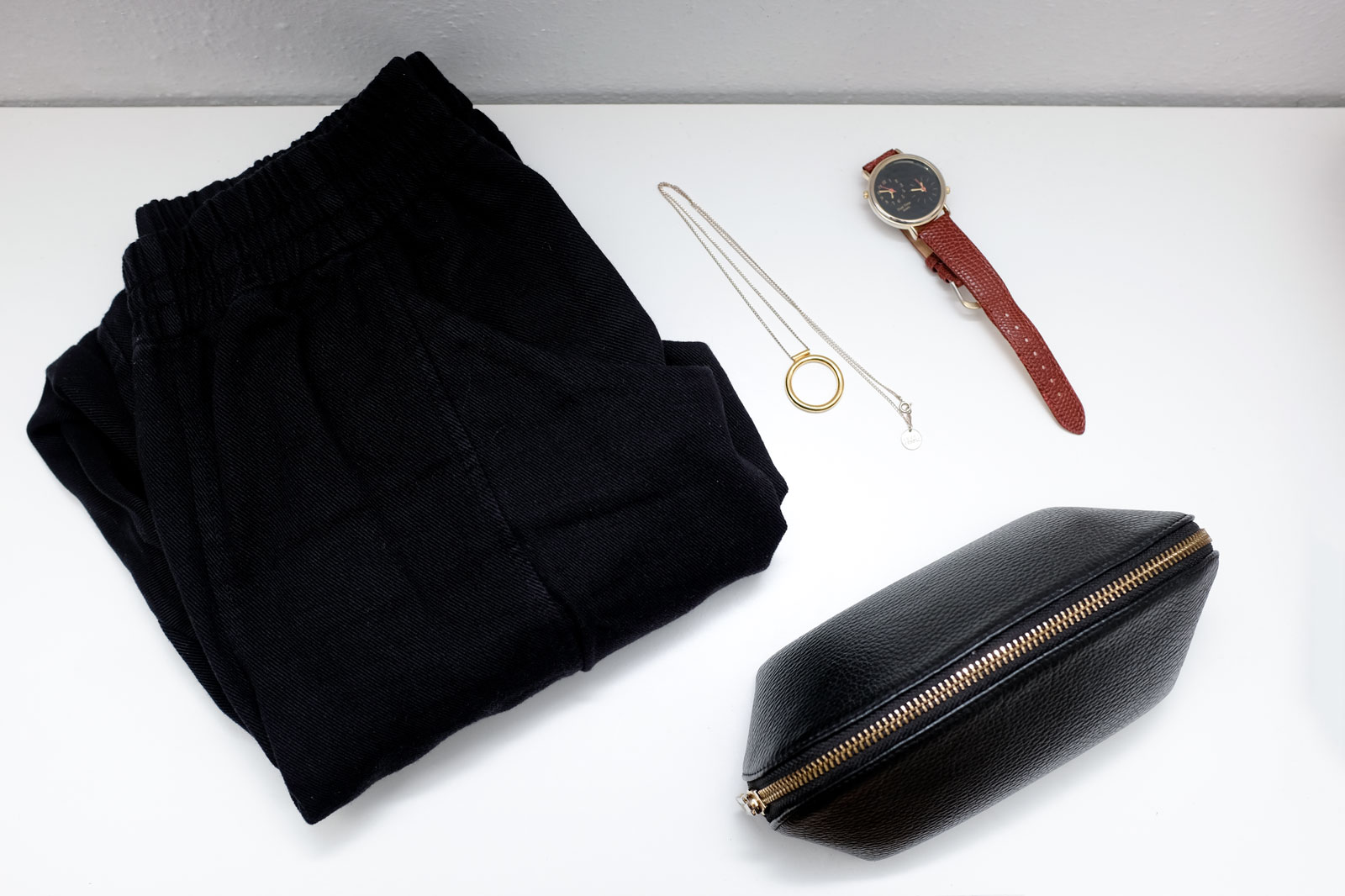 Pants, a necklace, a watch, and a cosmetic bag in a flatlay