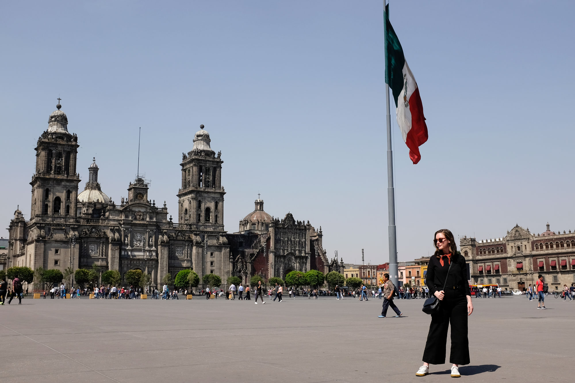 Alyssa standing in the Zocalo wearing a black long sleeve shirt, black wide leg pants, a rust scarf, and white sneakers