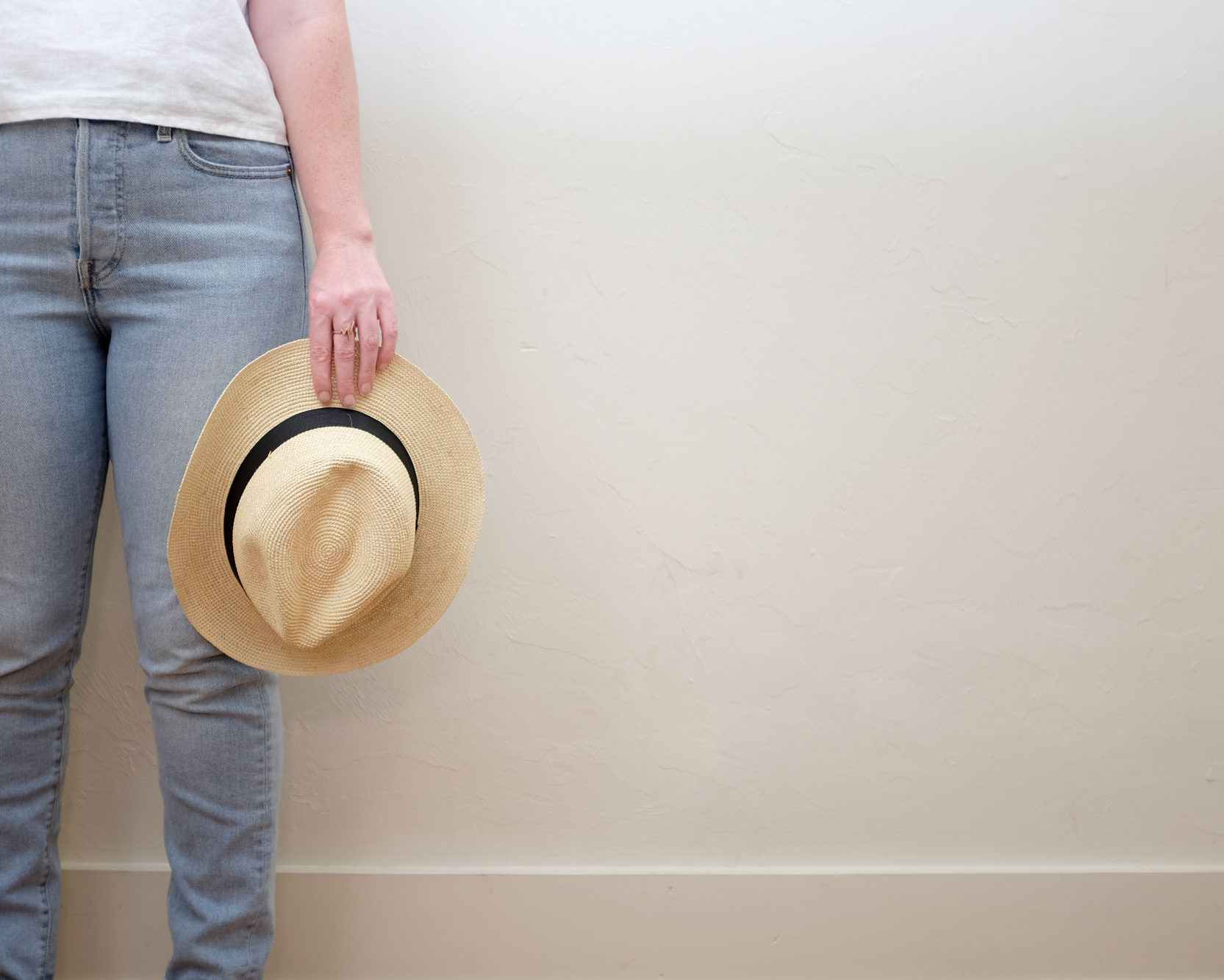 Krystal is wearing a white linen tee and light blue jeans and is holding a straw hat. The image is closely cropped on her hand and legs.