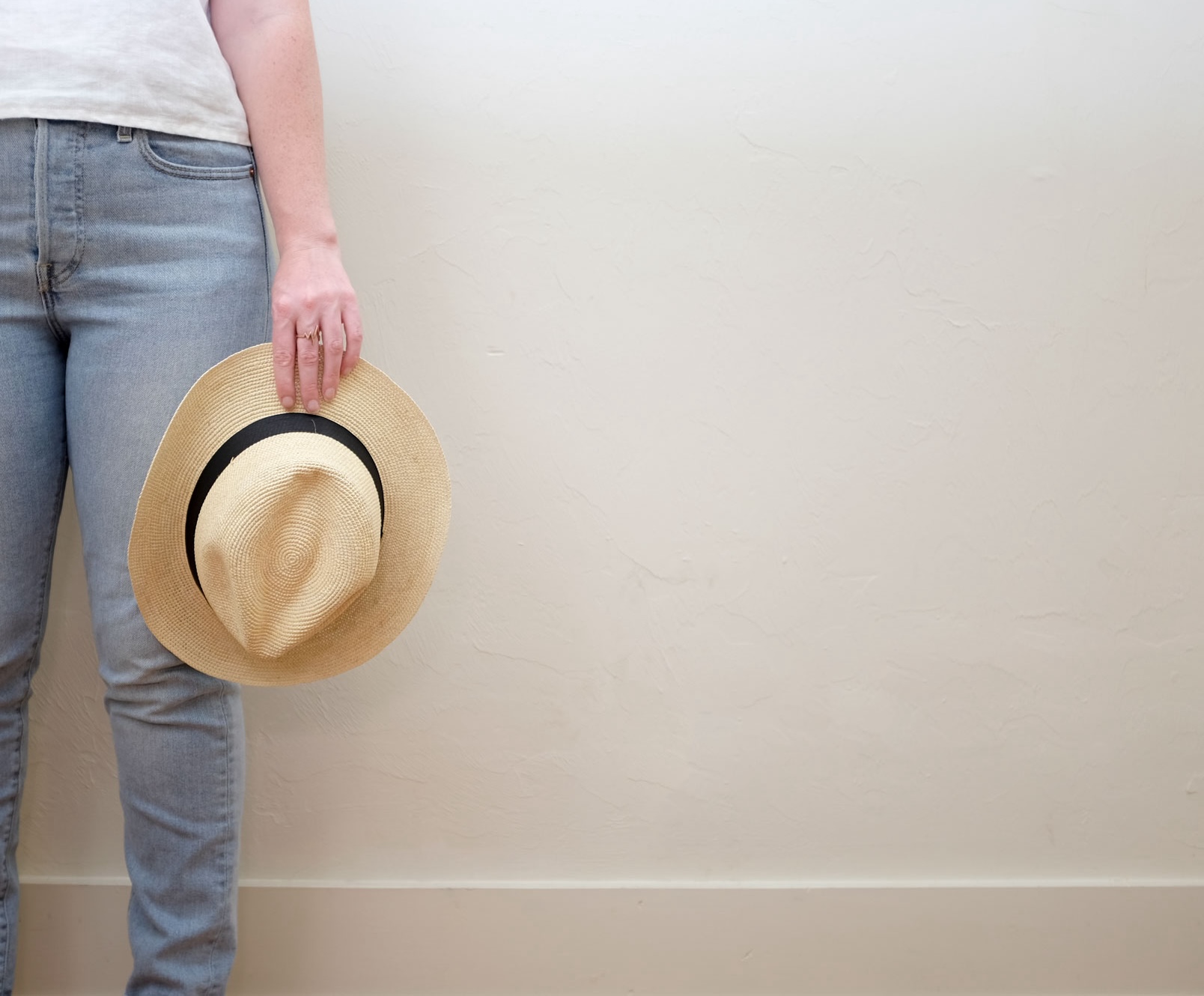 Alyssa is wearing a white linen tee and light blue jeans and is holding a straw hat. The image is closely cropped on her hand and legs.