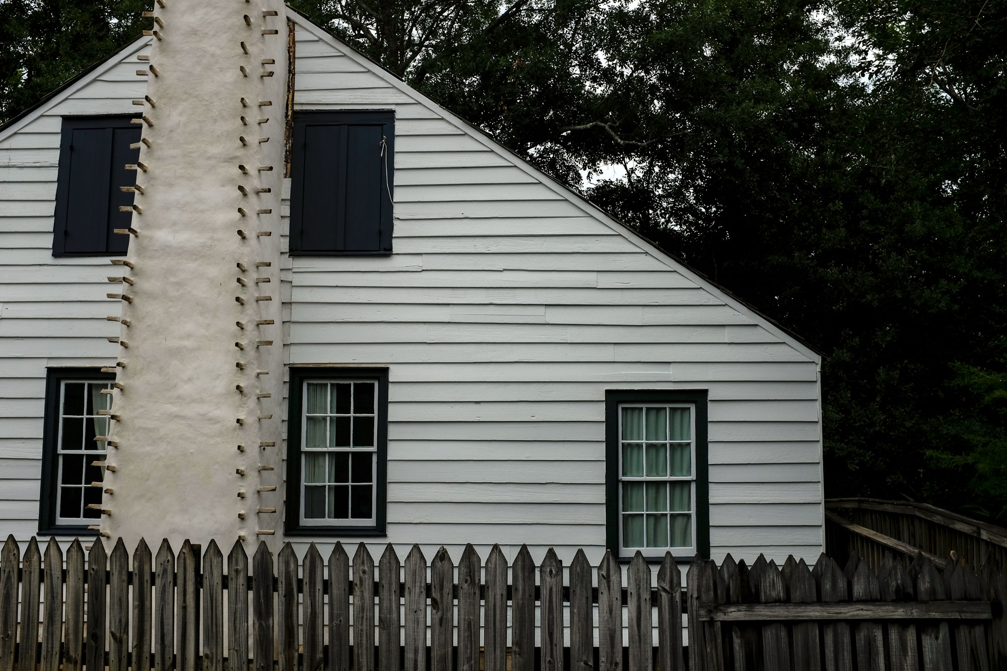 Side of a house at vermilionville. It is a white wooden house with a fireplace along the side and a wooden picket fence