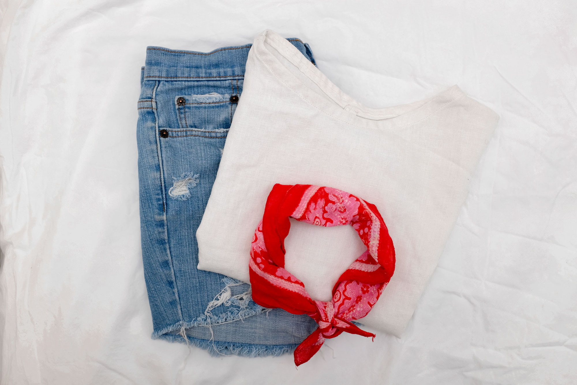 Flatlay with light wash denim cutoffs, a white linen top, and a red bandana