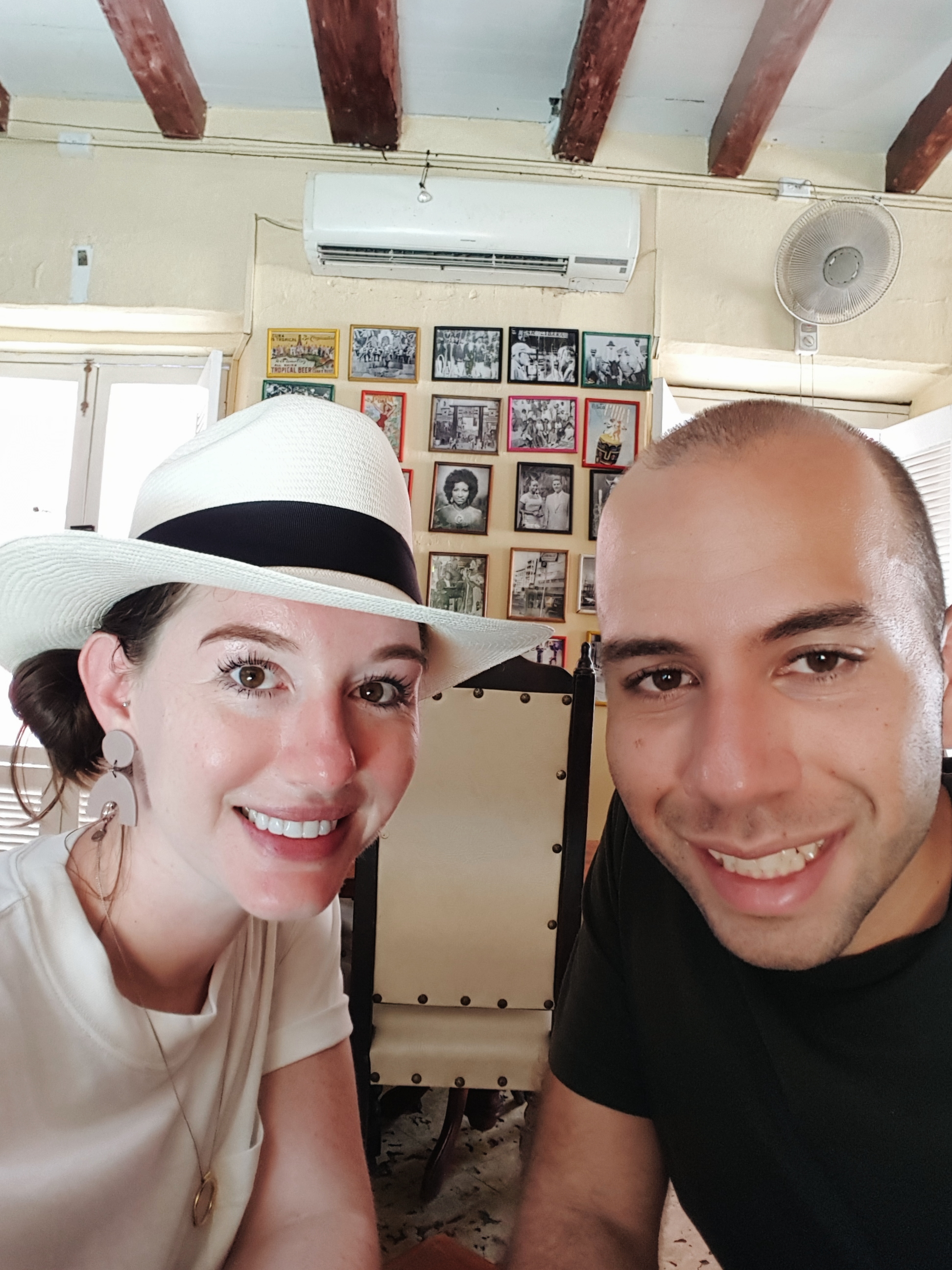 A selfie of Alyssa and Michael in a bar in Cartagena, Colombia. Alyssa is wearing a Panama hat and white tee, and Michael is wearing a black tee. They are both sweaty but smiling.