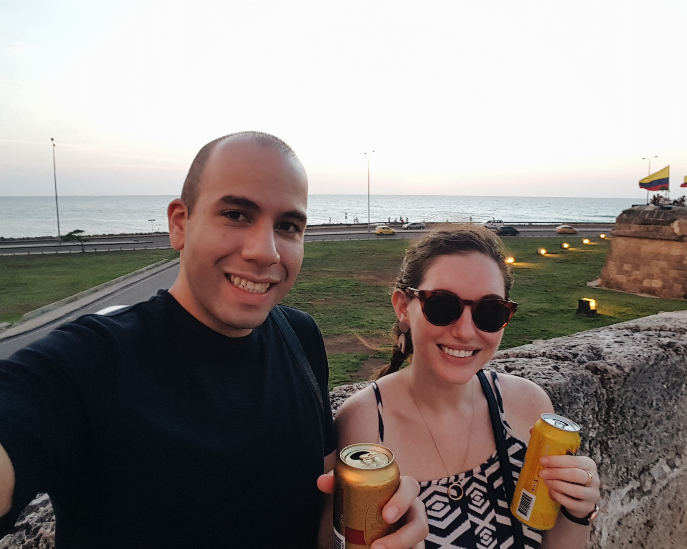 Krystal and Michael waiting for the sunset along the walls of the city, and they are each holding a beer. Michael is wearing a dark tee and Krystal is wearing a patterned dress and sunglasses