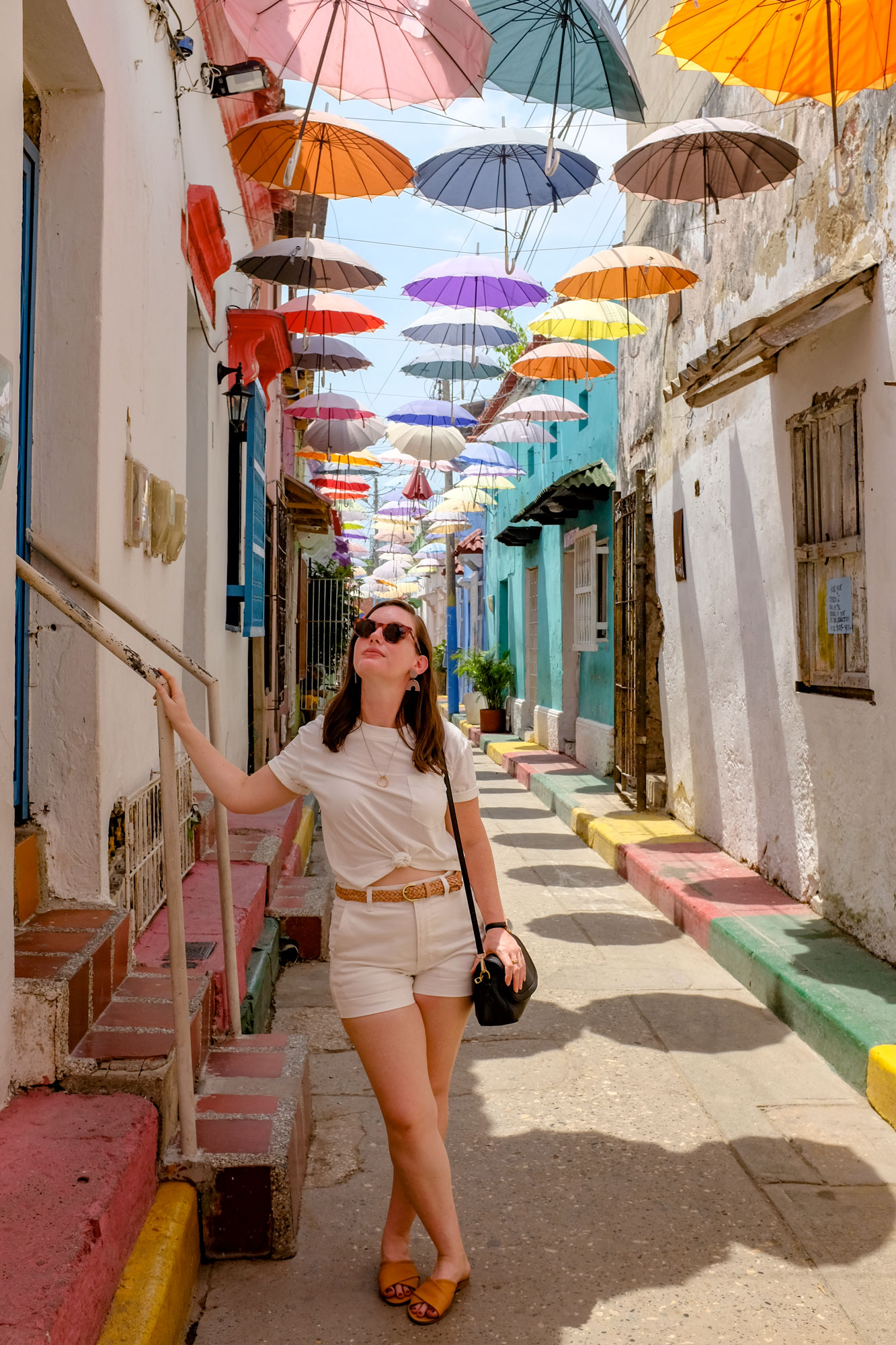 Alyssa standing under the colorful umbrellas on a street in Getsemani. She is wearing a white tee, white shorts, purse, belt and glasses