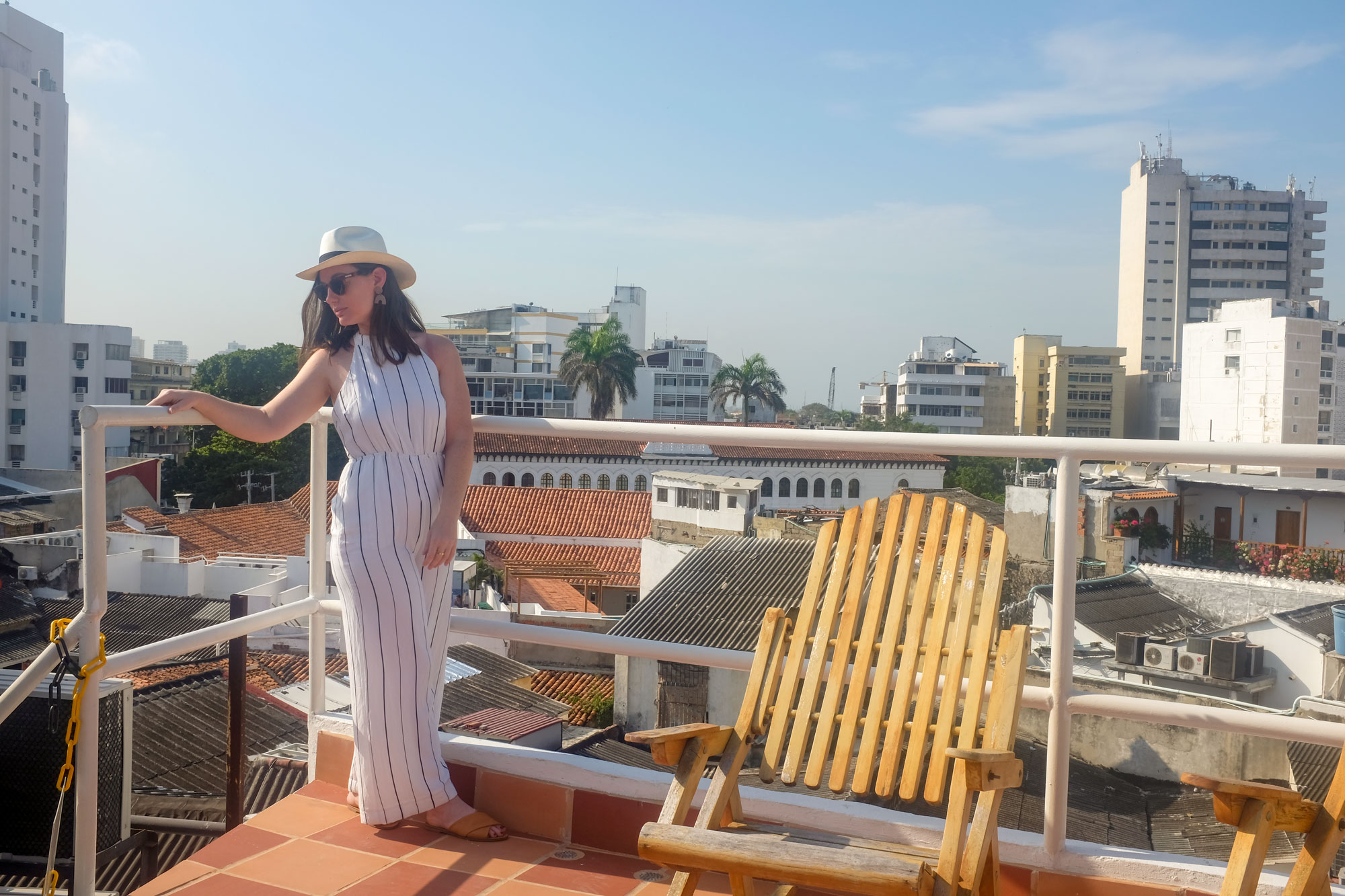 Krystal on the roof deck overlooking the city. She is wearing a white and blue striped jumpsuit, with a Panama hat, sunglasses, and brown sandals
