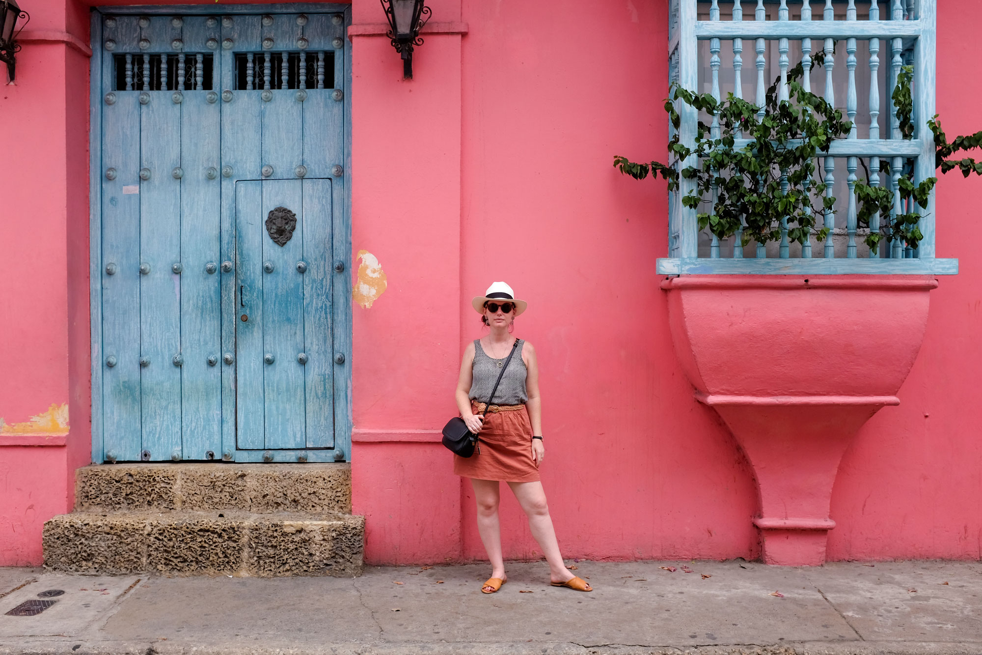 Krystal standing in front of a bright pink wall, looking at the camera. She is wearing a Panama hat, sunglasses, silk tank top, rust skirt, camel-toned sandals, and a black purse
