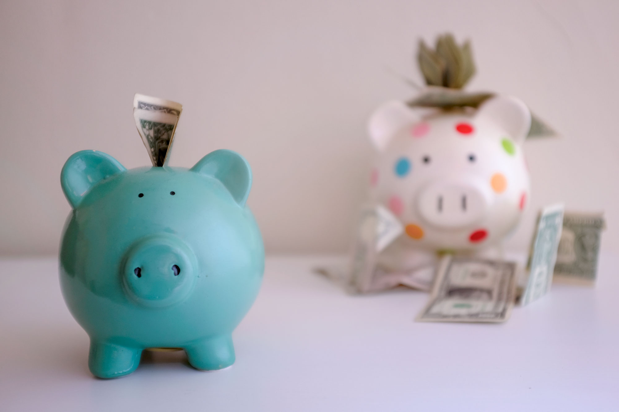 Two piggy banks. The small one is in the foreground and it just has a few dollars in the hole. The large piggy bank in the back has a lot of money in and around it.