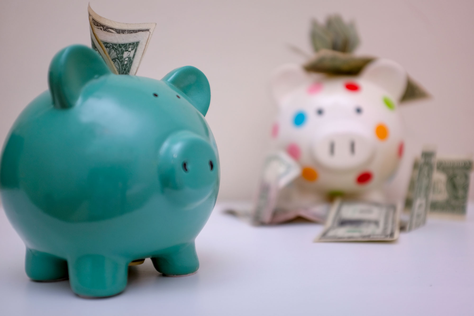 Two piggy banks. The small one is in the foreground and it just has a few dollars in the hole. The large piggy bank in the back has a lot of money in and around it.