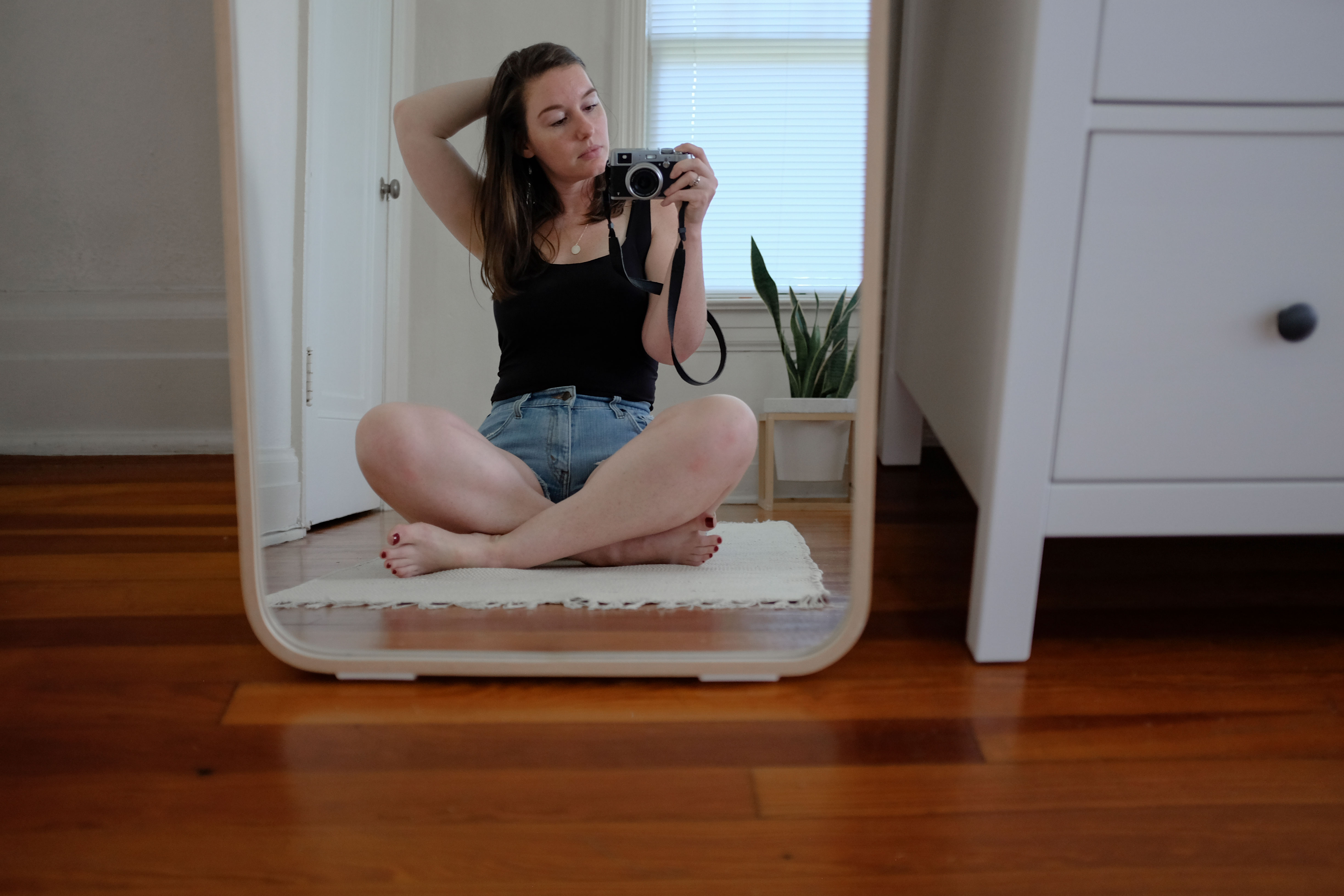 Alyssa sitting cross-legged in front of a mirror in a black tank and jean shorts
