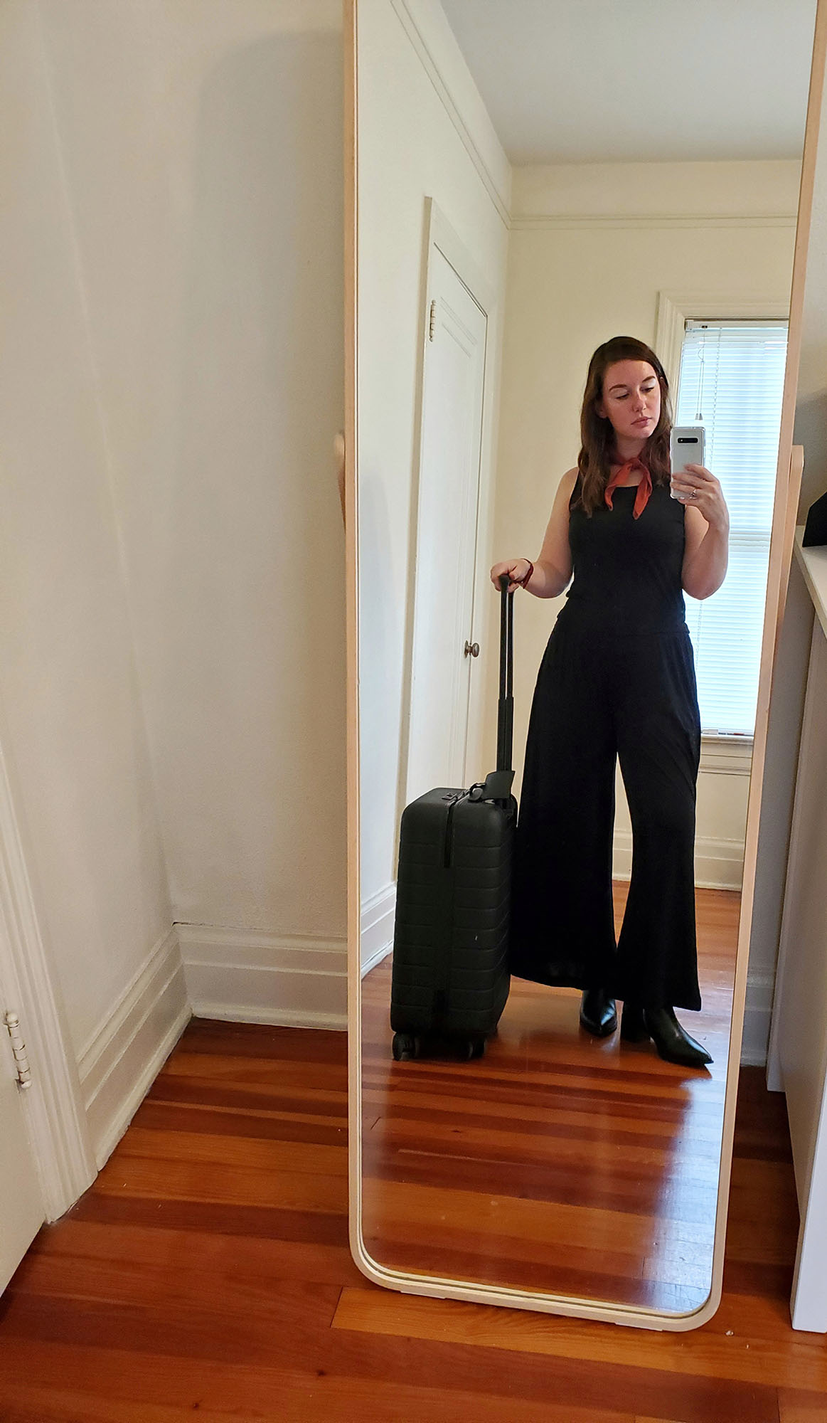 Krystal taking a photo in the mirror before her trip. She is wearing a black tank, black wide leg pants, black boots, and a red bandana. In her right hand is her carry on suitcase.