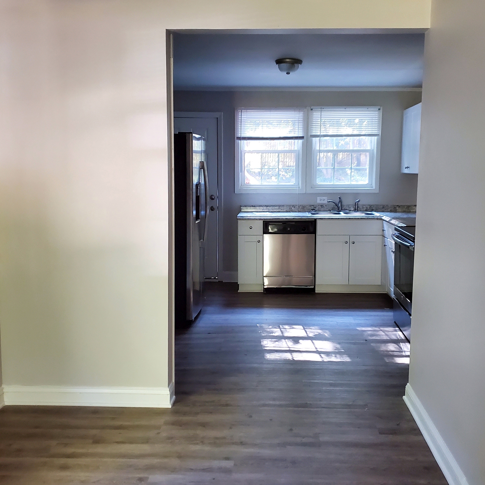 A view of a kitchen from another room; the dishwasher, fridge, sink, and oven are all visible and there is a window over the sink