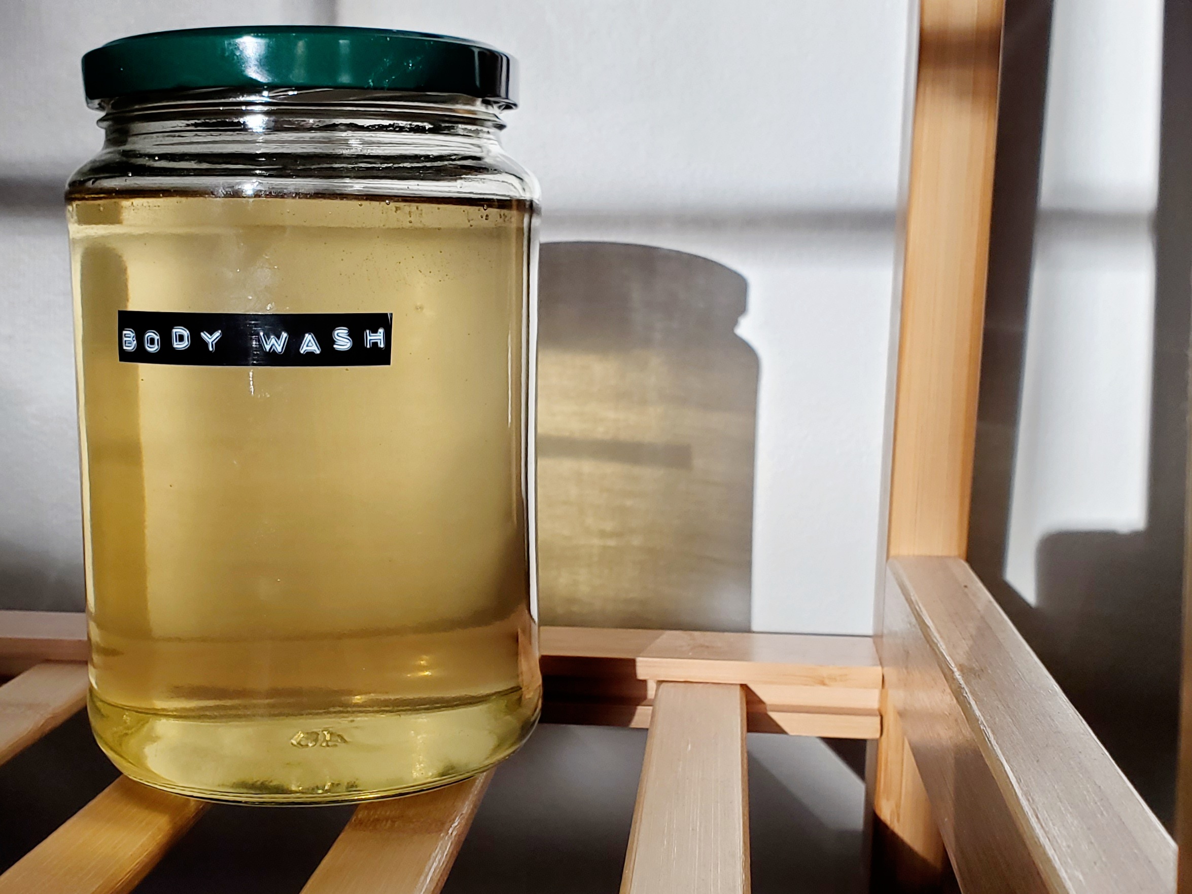 A large jar of pale yellow body wash with a green lid sits on a wooden rack. The sunlight is casting lots of shadows. The jar reads "body wash"