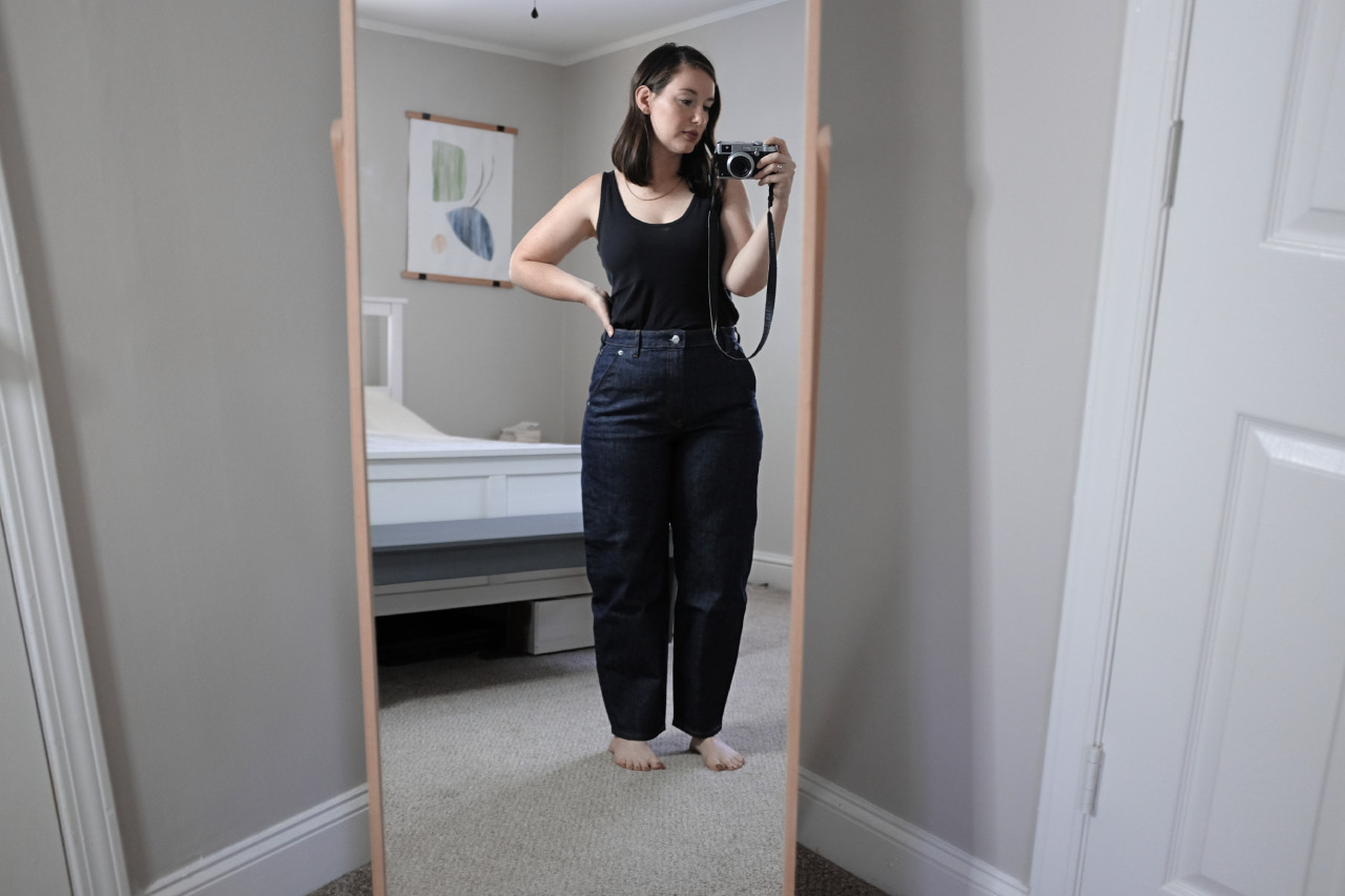 Alyssa is holding a camera and taking a photo in front of a full length mirror. She is wearing blue jeans and a black tank top.