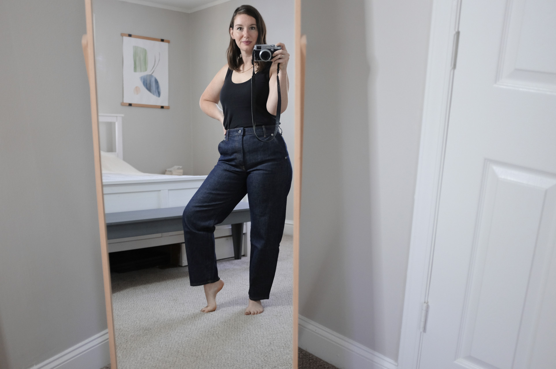 Alyssa holds a camera and takes a photo in the mirror while wearing dark blue jeans and a black tank