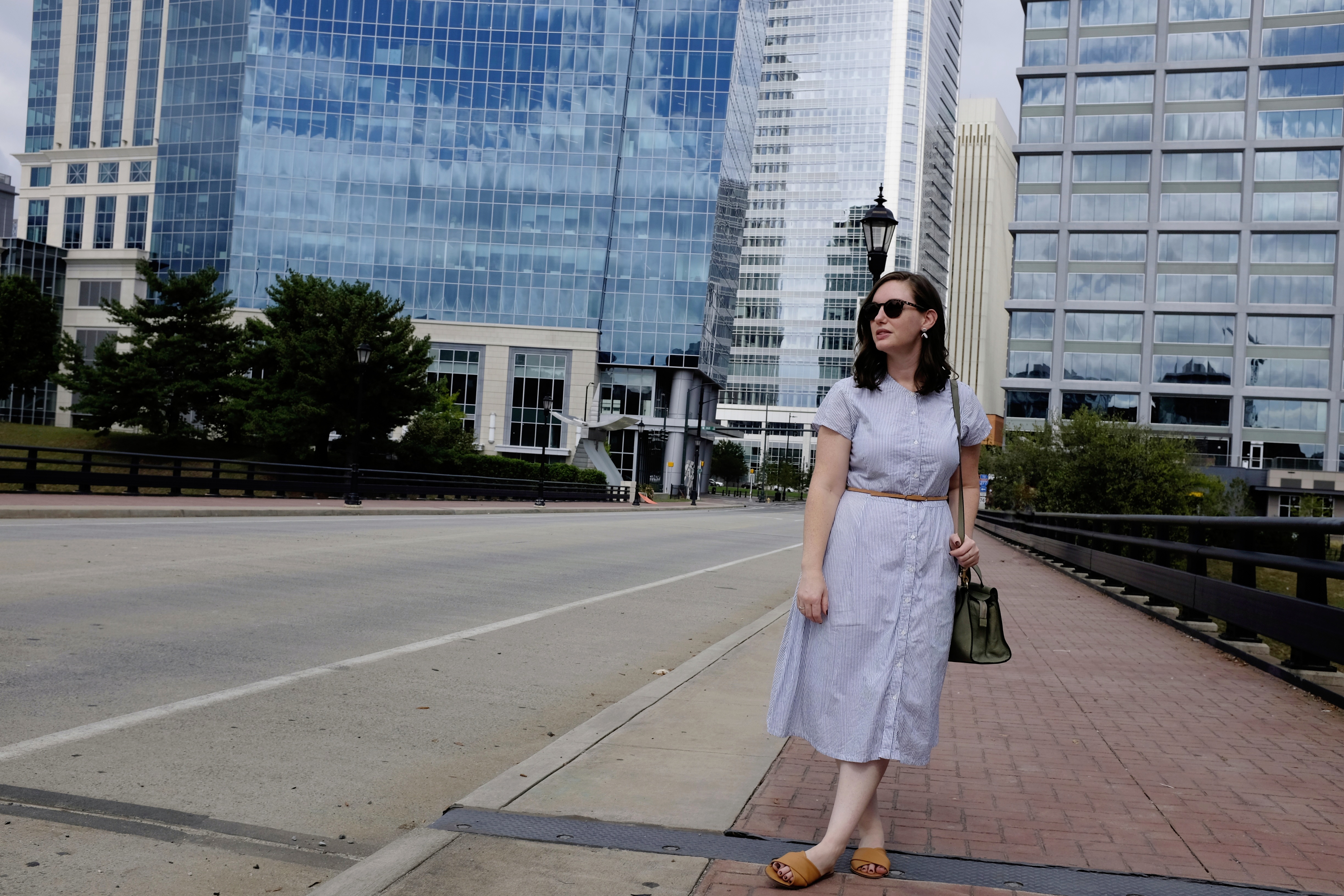 Alyssa stands in front of a street in the downtown area of Charlotte. You can see large buildings in the background, and she is wearing a blue striped dress with orange/brown sandals.