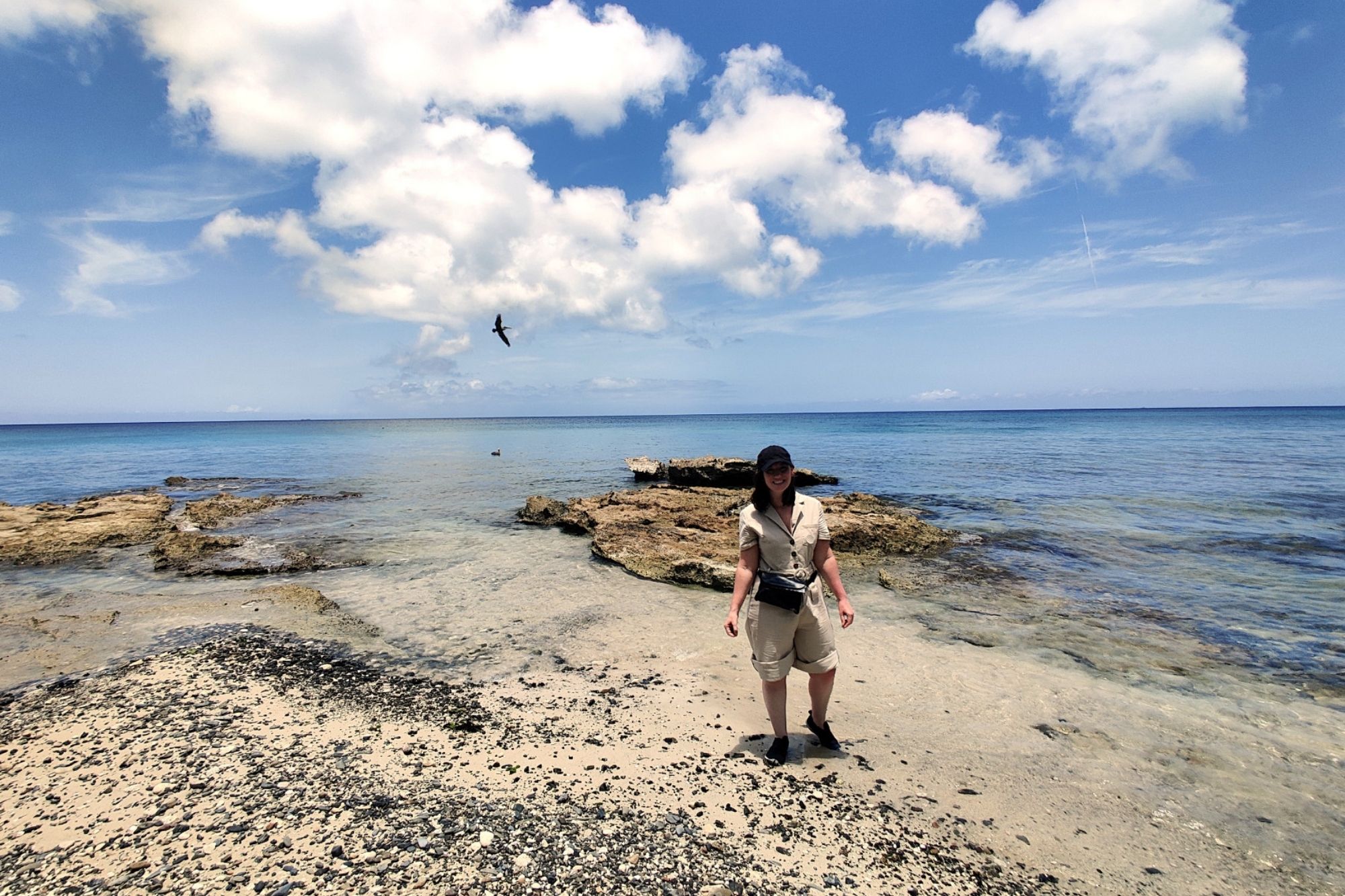 Alyssa stands in front of a rocky beach wearing a tan jumpsuit rolled to the knees, sneakers, a hat, and a belt bag