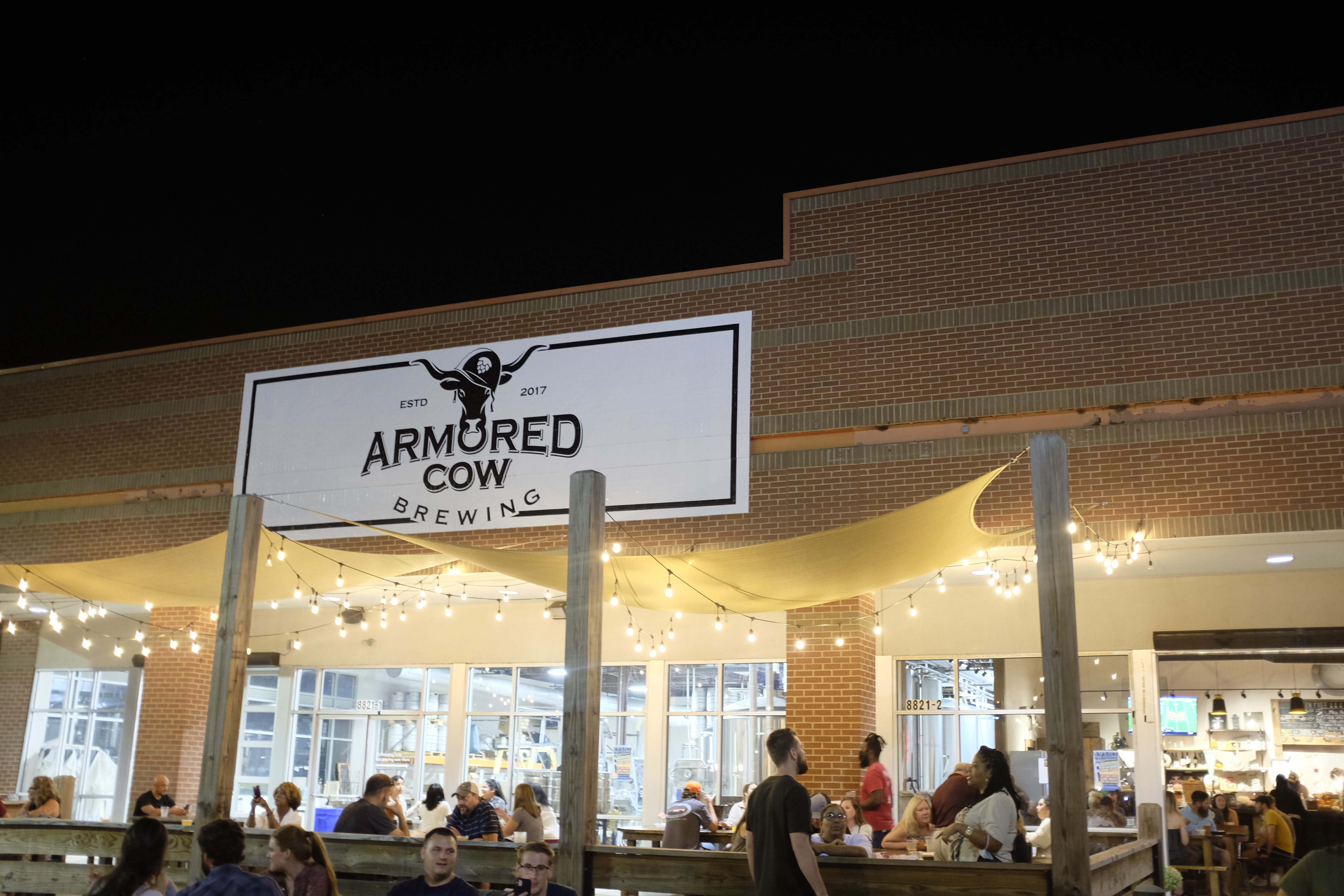 Exterior of Armored Cow Brewing; there are people sitting under canopies with string lights in front of a brick building.