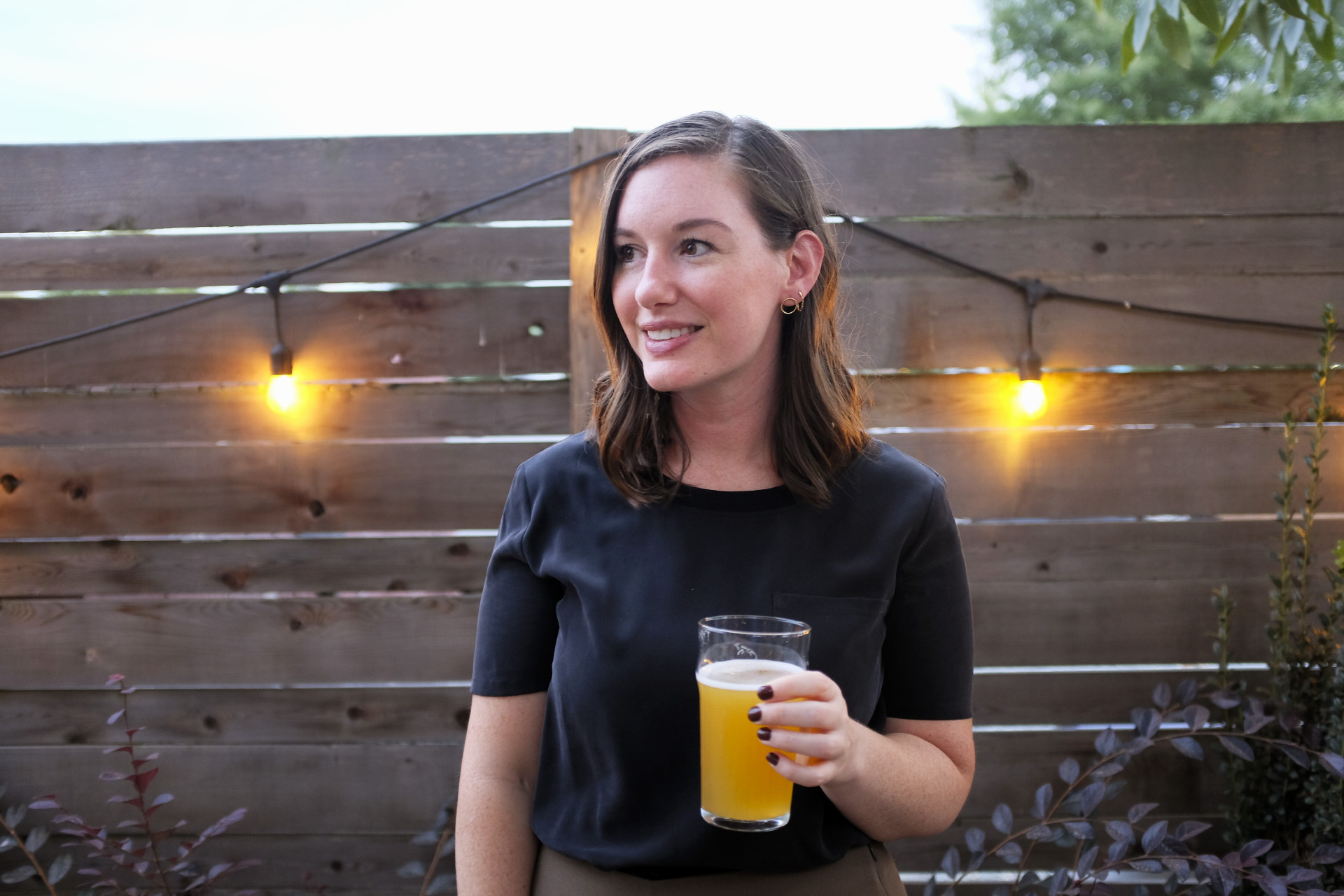 Krystal is standing in front of a wooden fence with string lights hung. She is wearing a black silk tee and holding a golden, hazy beer and smiling while looking left.