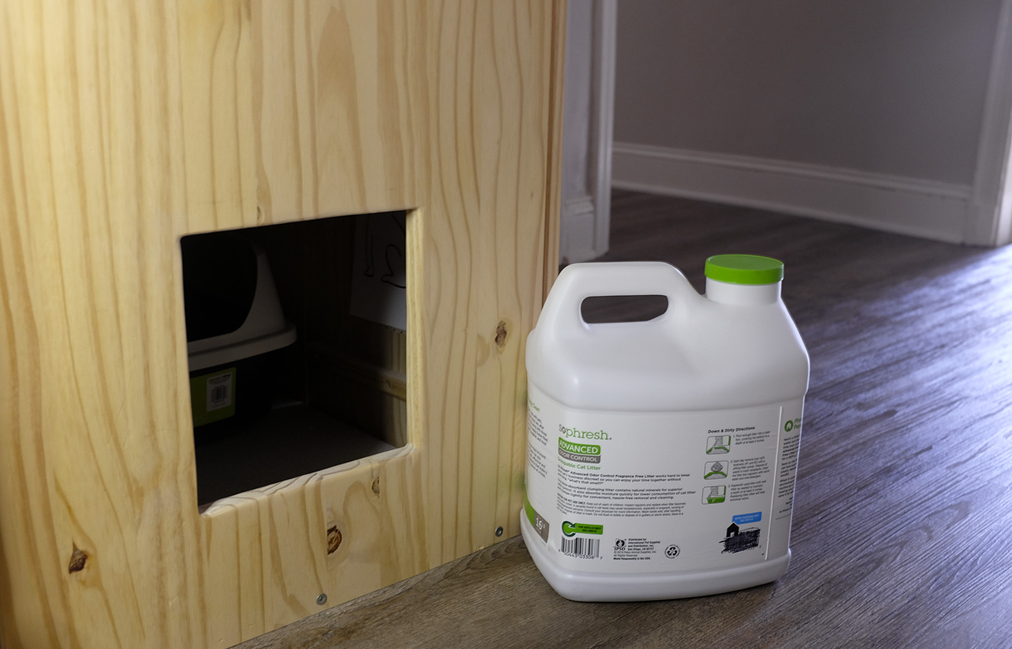 A wooden cabinet in a hallway has a square cutout, and you can see a litter box inside. In the foreground is a white kitty litter container 