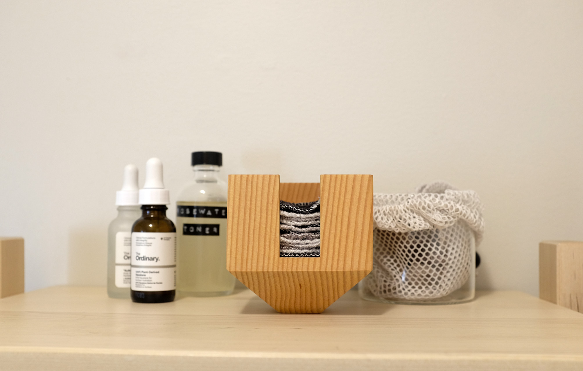 A bathroom shelf topped with several glass bottles of serums and toner, as well as a small wooden block containing cotton flannel face rounds and a glass container lined with a mesh bag