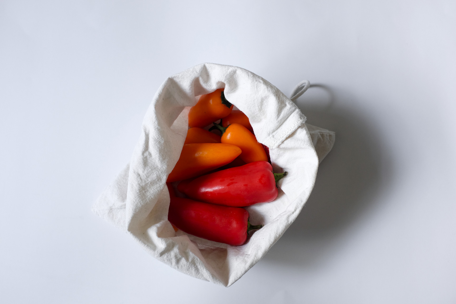 Red and orange mini sweet peppers sit in a cloth drawstring bag on a white surface