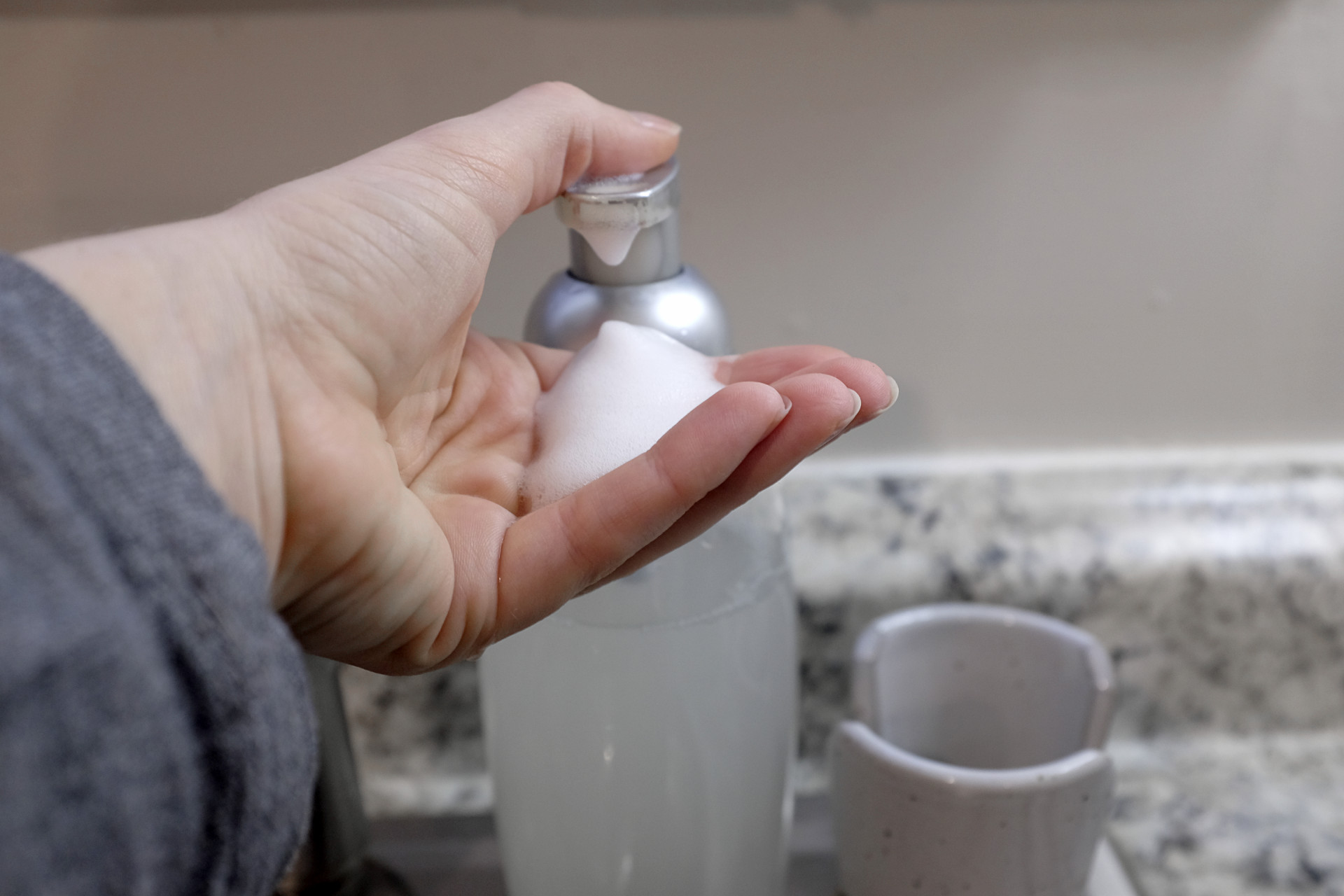 A caucasian hand pumps soap from a glass soap canister, and it's very foamy. The edge of a grey sleeve is visible