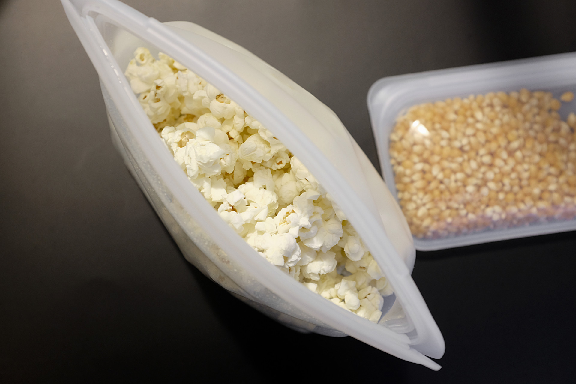 Two Stasher bags on a black surface. The larger one contains popped popcorn, and the smaller one contains unpopped kernels
