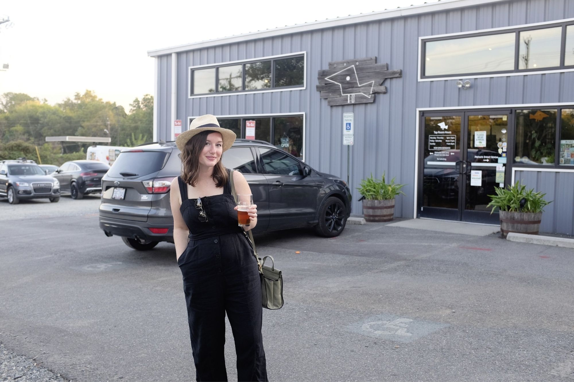 Alyssa is standing in front of Birdsong Brewing wearing a black tank jumpsuit and a Panama hat, holding a beer