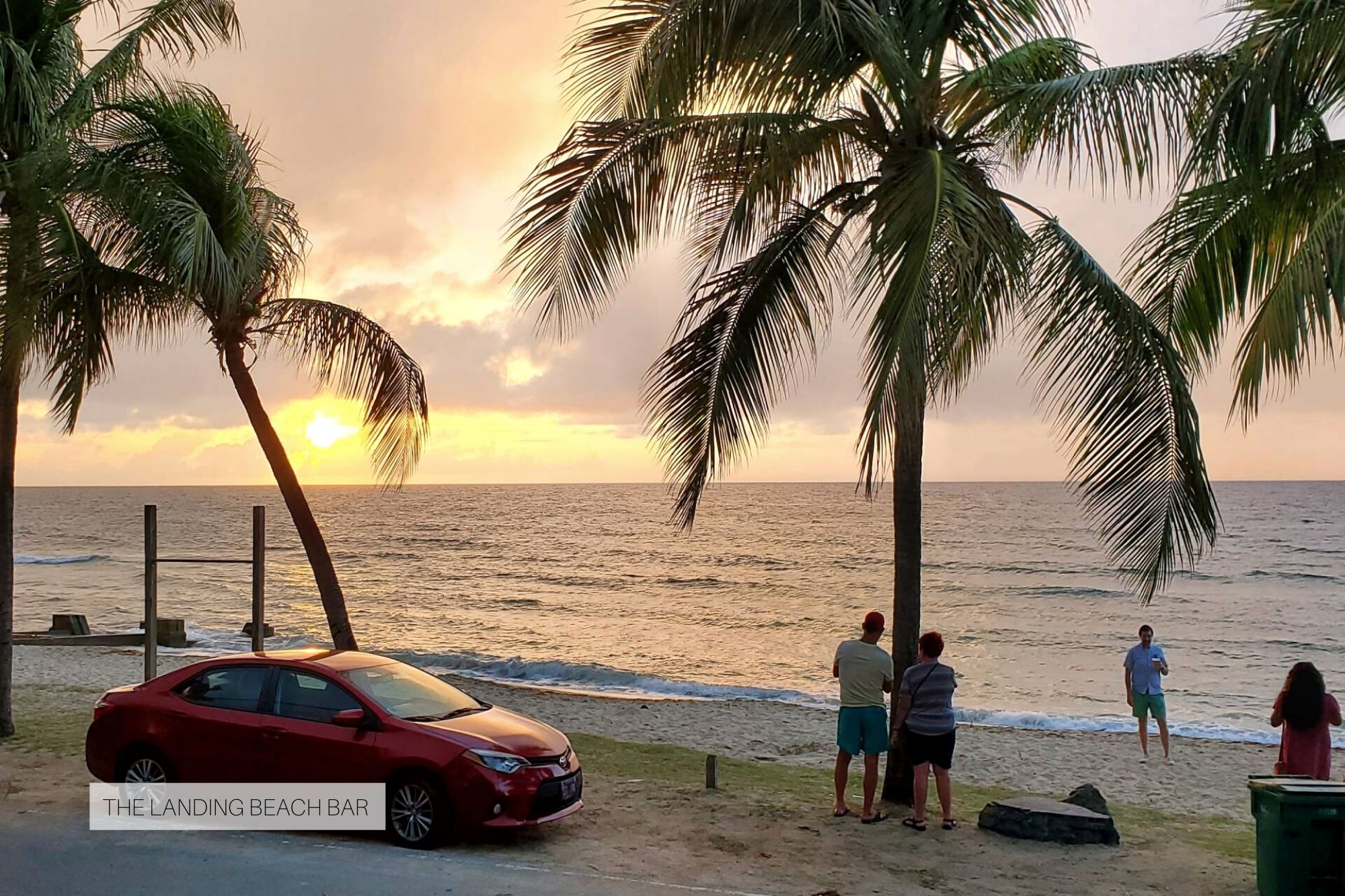 View from The Landing Bar - the sun is setting over the water, palm trees are in the foreground, and four people are watching or taking photos