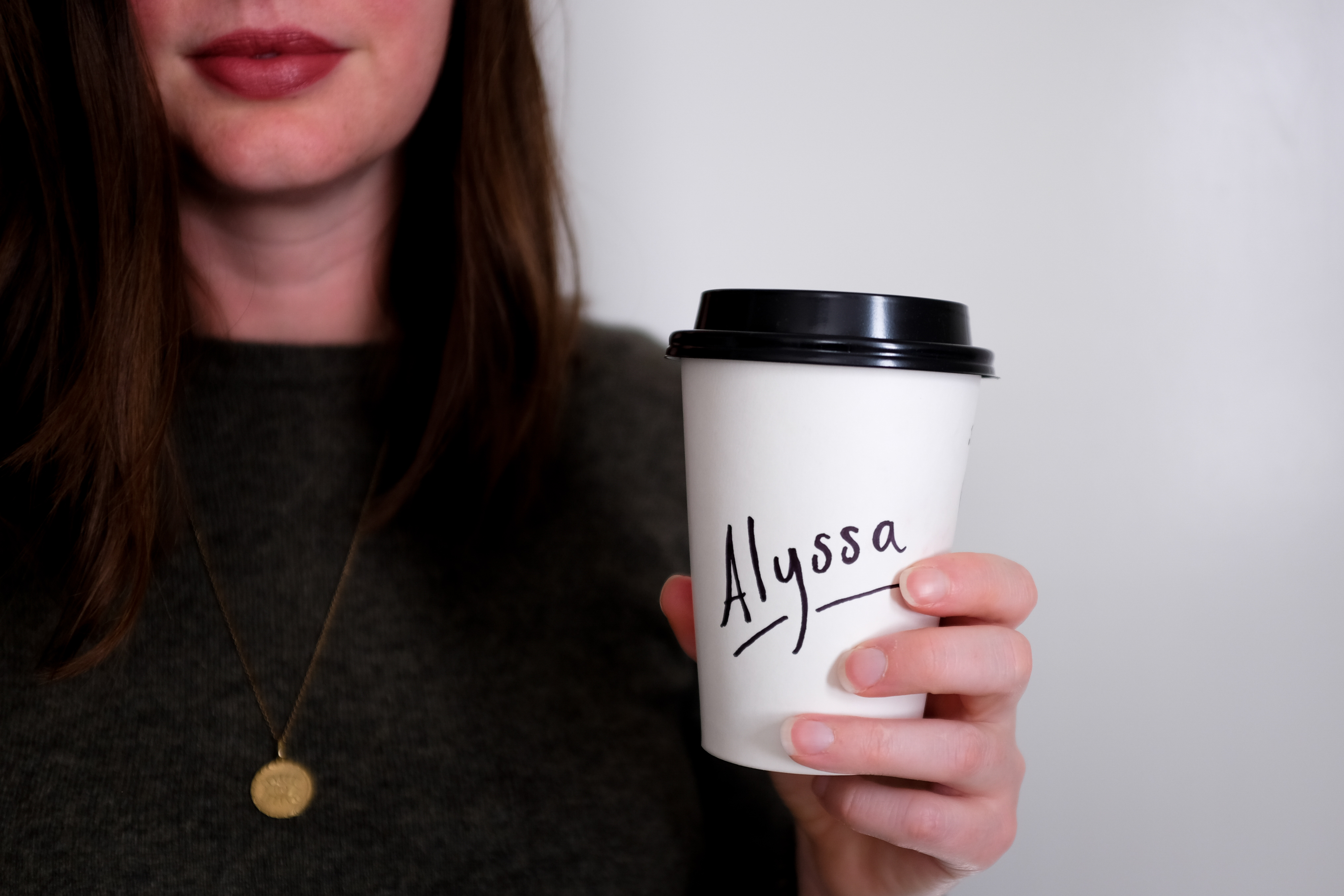 A white woman is wearing a green sweater and gold coin necklace and is holding a disposable coffee cup that reads "Alyssa." The image is very closely cropped to her face and upper body